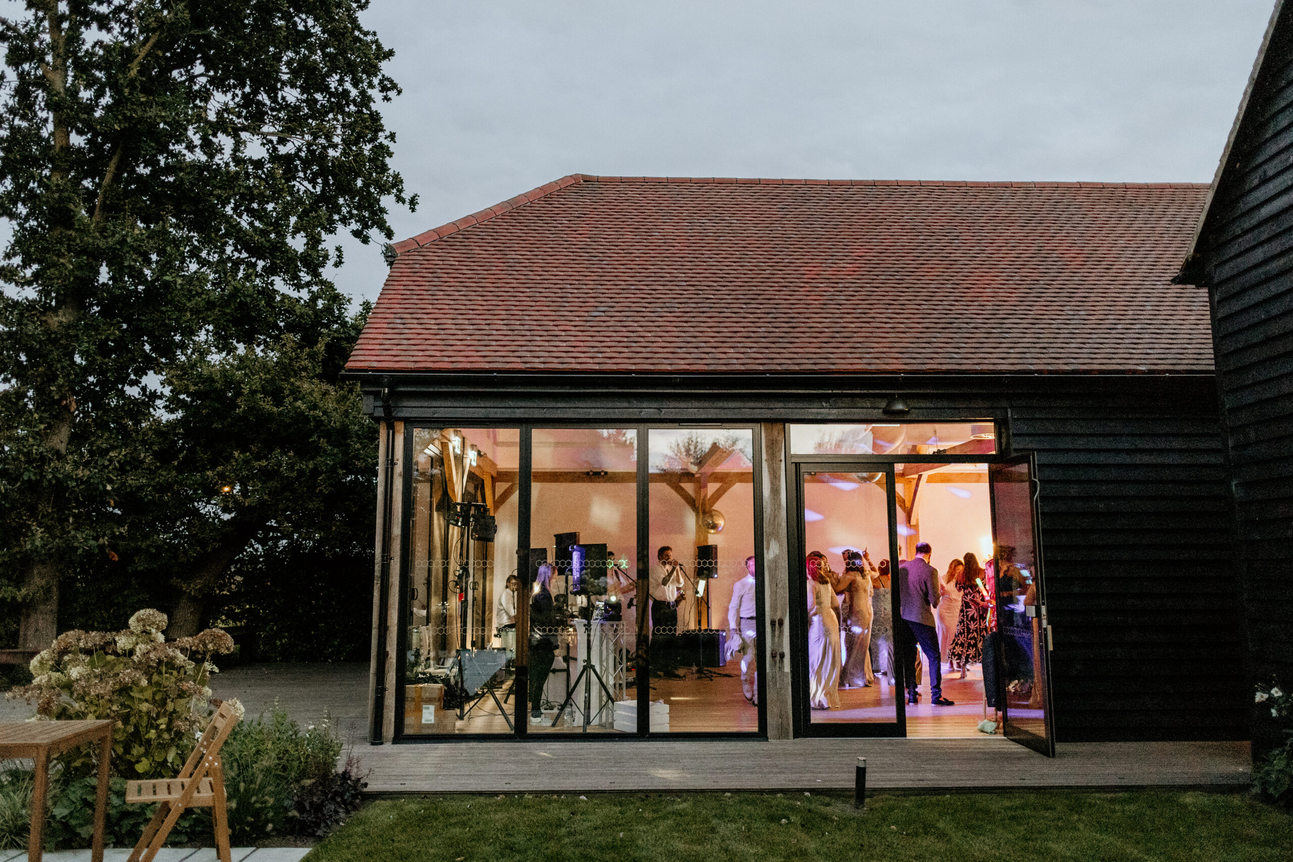 Exterior of Barn Wedding Venue with big glass windows. Inside guests are dancing.